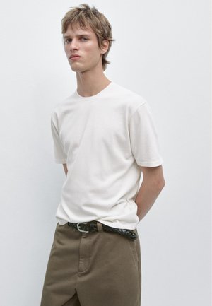 Giovane uomo in piedi contro una parete semplice, indossa una maglietta bianca, pantaloni marroni con cintura nera intrecciata, mani dietro la schiena, espressione neutra.