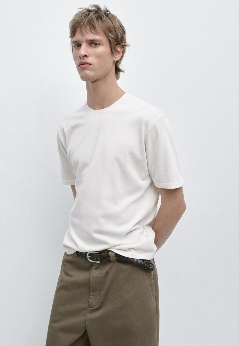 Jonge man staat tegen een effen muur, draagt een wit T-shirt, bruine broek met gevlochten zwarte riem, handen achter de rug, neutrale uitdrukking.