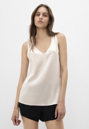 Kvinde står mod en ensfarvet hvid baggrund iført en beige satinærmeløs top og sorte shorts og kigger direkte ind i kameraet.