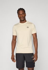 Μπεζ κοντομάνικη μπλούζα από μαλακό ύφασμα, με στρογγυλή λαιμόκοψη και μικρό μαύρο λογότυπο στο στήθος. Συνδυασμένη με σκούρα σορτς.