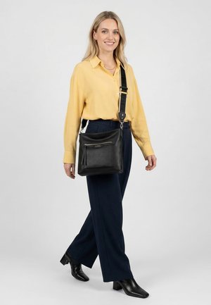 Naine kollases pluusis ja tumedates pükstes kõnnib, kandes musta nahast ristihoidmist laia rihmaga, naeratades ühevärvilise tausta ees.