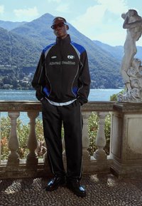 Czarny dres z niebieskimi akcentami i białymi lamówkami, z napisem "Altered Realities". Nosi się go z błyszczącymi czarnymi butami. Sceneria na świeżym powietrzu.