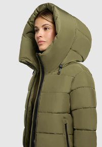 Olijfgroen pufferjack met een oversized capuchon, gewatteerde textuur, ritssluiting aan de voorkant en zwarte ritsdetails. Heeft zijzakken.