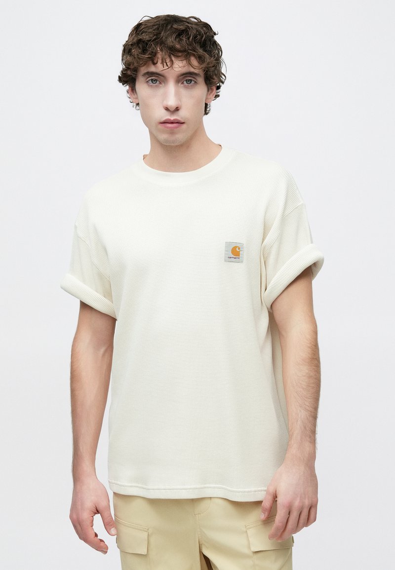 Light beige textured t-shirt with rolled sleeves and a square logo patch on the chest. Relaxed fit, soft fabric, and crew neckline.