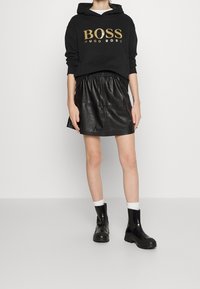 Sudadera negra con capucha y estampado dorado "BOSS HUGO BOSS", combinada con falda de cuero negra y botines negros. Medias blancas visibles.