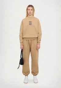 Beiger Pullover mit dem Schriftzug "BODY MIND SOUL", kombiniert mit passenden Jogginghosen. Weicher Stoff, lockere Passform und Bündchen an den Knöcheln. Dazu weiße Sneakers getragen.