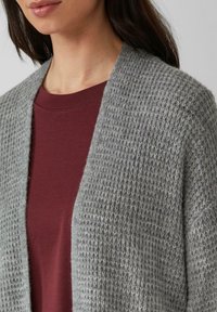 Hellgraue Strickjacke mit strukturierter Musterung, offene Vorderseite, kombiniert mit einem bordeauxfarbenen T-Shirt darunter. Keine sichtbaren Knöpfe oder Verschlüsse.