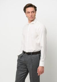 Camicia bianca a maniche lunghe con colletto classico, abbinata a pantaloni grigi con pieghe e una cintura in pelle marrone. Tessuto liscio, vestibilità su misura.