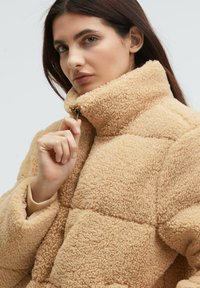 Beige, fluffy fleece jacket with a high collar and zip closure. It features a quilted design and textured surface, including large panels.