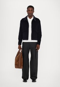 Marineblå fløjlsk jacket med frontlommer, hvid polo underlag, sorte brede bukser og brun ruskinds duffeltaske med læderdetaljer.