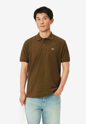 Hombre que lleva una camiseta tipo polo marrón de manga corta con logo de cocodrilo verde y vaqueros azul claro, de pie con una mano en el bolsillo y sosteniendo una tela blanca.