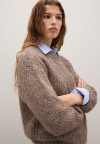 Lichtbruin mohair-blend sweater met een losse pasvorm en een gestructureerde brei, gedragen over een lichtblauw overhemd met omgeslagen mouwen.