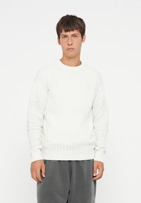 Hvid tekstureret sweater med horisontale ribber og diamantmønstre, med rund halsudskæring og ribbede manchetter. Båret med grå bukser.