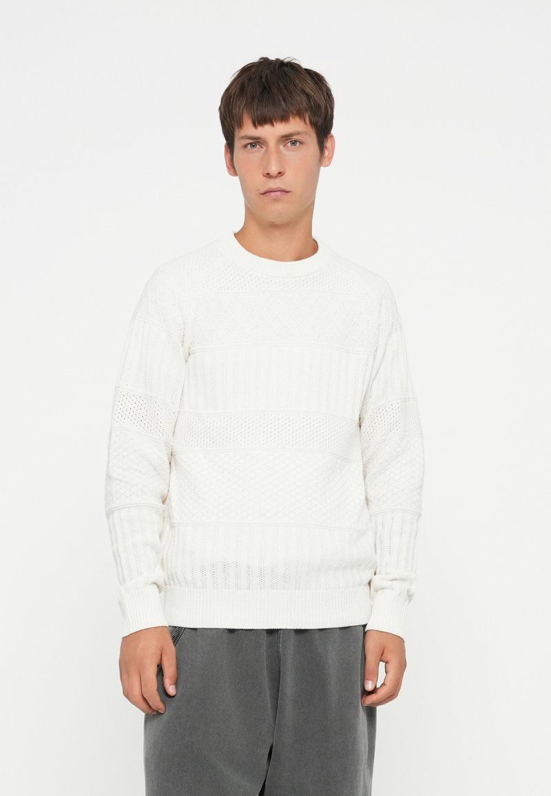 Hvid tekstureret sweater med horisontale ribber og diamantmønstre, med rund halsudskæring og ribbede manchetter. Båret med grå bukser.