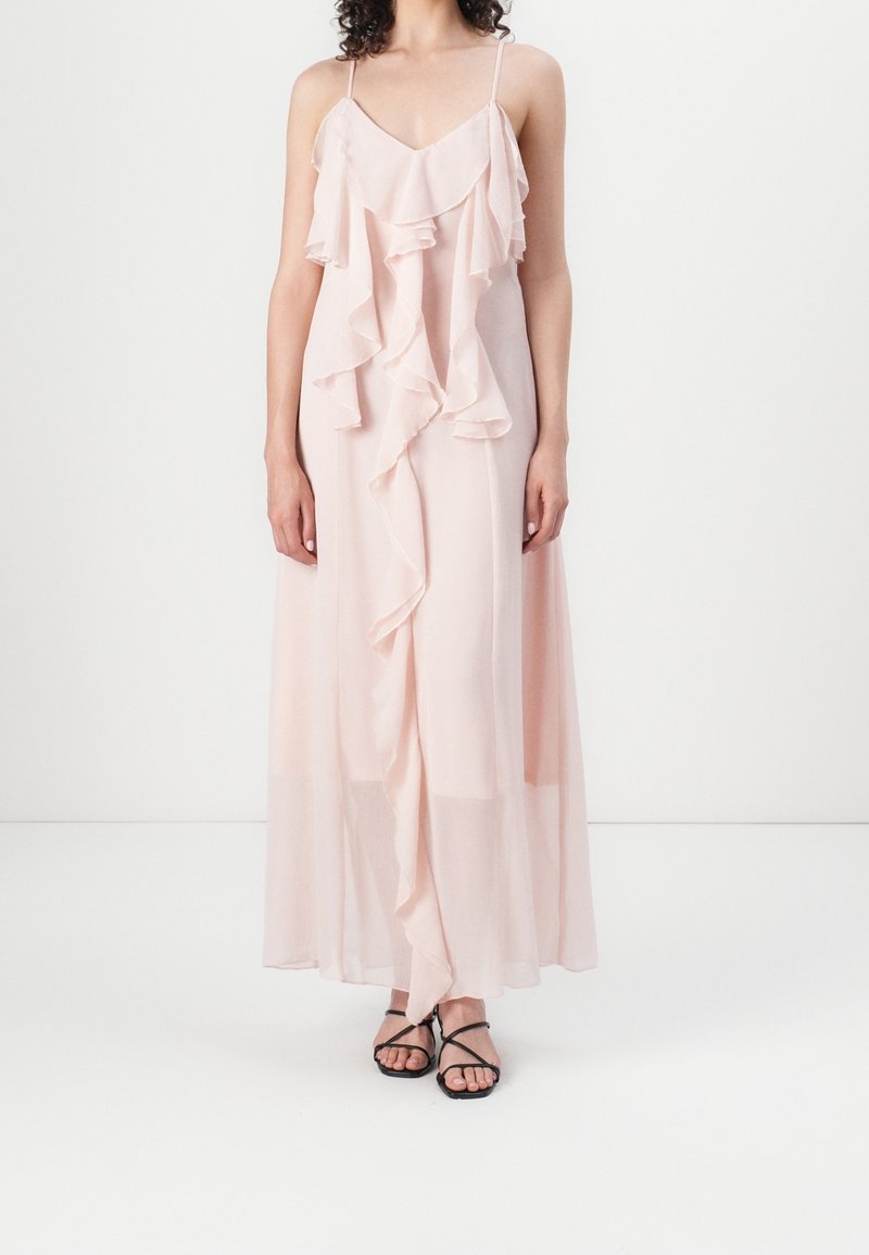 Robe longue rose clair avec détails à volants, bretelles spaghetti et texture fluide. Associée à des sandales noires à lanières.