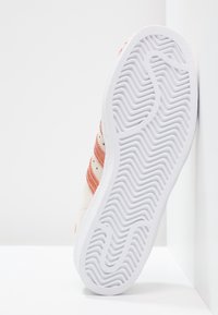 Witte sneaker met een gestructureerde zool, een zachte bovenkant met koraalkleurige strepen en perforaties, schuin geplaatst tegen een lichte achtergrond.