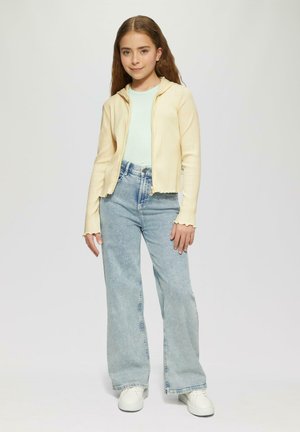 Lichtgele geribbelde ritshoodie, mintgroene top, high-waisted lichtblauwe flare jeans, witte sneakers en een gegolfde mouwdetail.