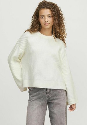 Pull en tricot côtelé de couleur crème, avec un col rond, des épaules basses et une coupe décontractée. Associé à un jean gris délavé.