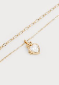 Złoty łańcuszek z naszyjnikiem w kształcie serca, zakończonym zawieszką o perłowym wykończeniu. Ogniwa łańcucha są wydłużone i owalne.