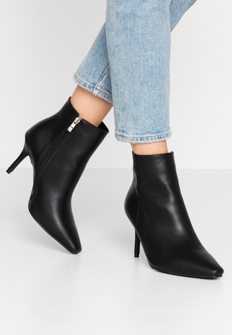 Schwarze spitz zulaufende Leder-Ankle-Boots mit seitlichen Reißverschlüssen, getragen mit hellblauen Cropped-Jeans auf einem weißen Boden.