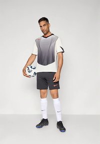 Vaaleanharmaa ja mustakuvioinen urheilupaita lyhyillä hihoilla, yhdistettynä mustiin shortseihin, valkoisiin sukkiin ja mustiin nappulakenkiin; pitelee jalkapalloa.