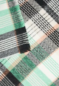 Tissu tissé à carreaux présentant des teintes de vert, noir, blanc et orange. Les motifs distincts incluent des rayures et des zigzags avec des bords frangés.