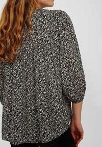 Blouse noire avec un motif floral crème, coupe ample, manches bouffantes et dos froncé. Convient pour un usage décontracté.