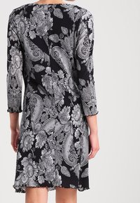 Czarna sukienka z białymi misternymi wzorami kwiatowymi i paisley, z rękawami do trzech czwartek i lekko rozszerzanym dołem, pokazana z tyłu.