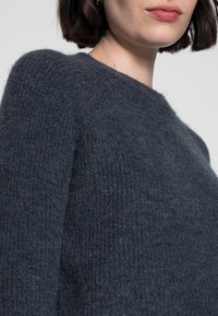 Pull tricoté gris foncé avec une surface texturée et un col rond classique, doté de manches légèrement bouffantes pour plus de forme.
