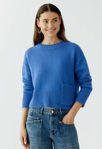 Gekürzter blauer Pullover aus weichem Strick mit geripptem Rundhalsausschnitt und Bündchen, ausgestattet mit einer kleinen Patttasche auf der Vorderseite. Kombiniert mit Jeans.