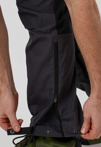 Mains ajustant le bouton-pression et la fermeture éclair au bas des jambes d’un pantalon de plein air sombre sur des chaussures de randonnée vertes.