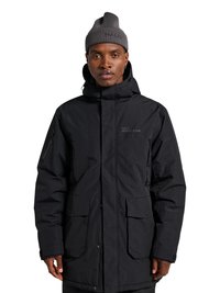 Mies pukeutuneena mustaan hupulliseen talvitakkiin, jossa isot etutaskut, ja harmaaseen neulottuun pipoon, katsoo suoraan neutraalilla ilmeellä.