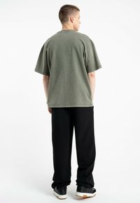 Przestronna oliwkowozielona bawełniana koszulka z krótkimi rękawami, okrągłym dekoltem i widocznymi szwami, zestawiona z czarnymi luźnymi spodniami i czarnymi trampkami.