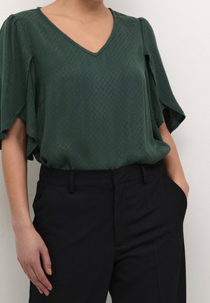 Woman wearing a dark green textured blouse with flutter sleeves and black tailored pants with one hand in pocket.