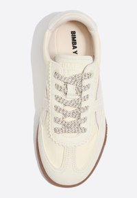 Kremasto obarvan športni čevelj z vzorčastimi vezalkami, šivanim detajlom in blagovno znamko "BIMBA Y LOLA" na jezičku in vložku, viden od zgoraj.