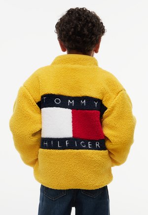 Dieťa s kučeravými vlasmi oblečené v žltej fleecovej bunde s veľkým logom Tommy Hilfiger na chrbte, stojace s rukami v vreckách.