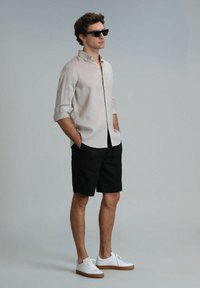 Chemise en lin claire à boutons et aux manches retroussées, associée à un short noir et des baskets blanches. Le mannequin porte des lunettes de soleil noires.