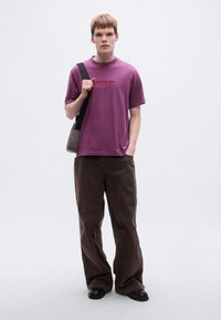 Μωβ βαμβακερό μπλουζάκι με κείμενο στο μπροστινό μέρος, συνδυασμένο με χαλαρά καφέ παντελόνια από κοτλέ. Το μοντέλο κρατά μια γκρι τσάντα και φοράει μαύρα παπούτσια.