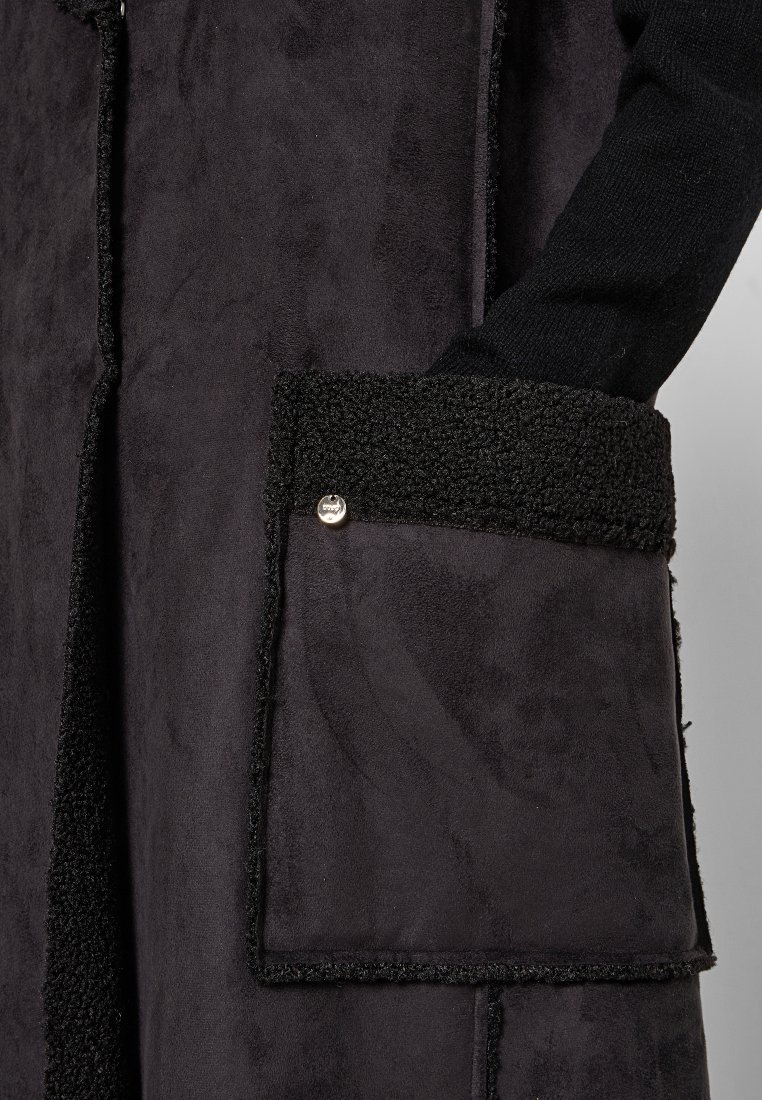 Schwarzer Wildleder-Mantel mit einer strukturierten schwarzen Stofftasche, ausgestattet mit einem silbernen Druckknopf und kontrastierenden Fleece-Details entlang der Kanten.