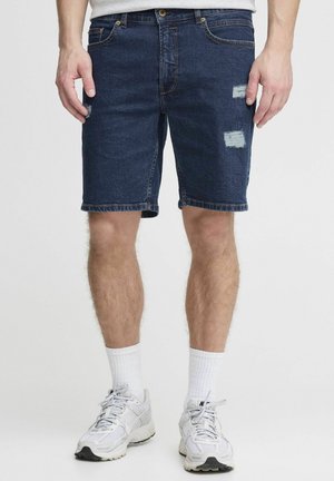 Dunkelblaue Jeansshorts mit lockerem Sitz. Verfügt über ausgefranste Stellen und einen geraden Saum. Getragen mit weißen Sportsocken und grauen Turnschuhen.