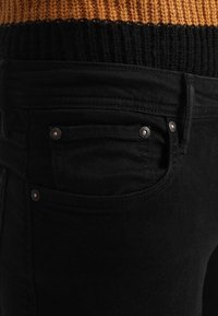 Musta denimi-farkut, joissa lähikuva sivutaskusta, näkyvissä kupariset niitit, sileä pinta ja hienovaraiset ompelusidokset.