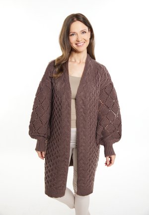 Bruine, gebreide cardigan met textuurpatronen en ballonmouwen. Open voorontwerp met geribde manchetten, gedragen over een lichte top en broek.
