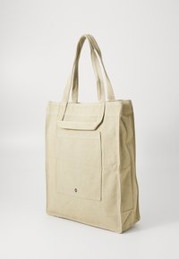 Beige canvas tote bag with two handles, a front pocket, and structured design. Features visible stitching and a small logo detail.