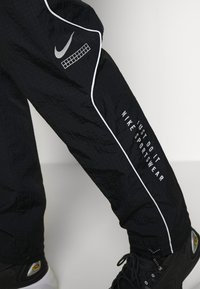 Svarta Nike sportbyxor med vit piping och texten "Just Do It Nike Sportswear" som bärs med svarta och vita sneakers.