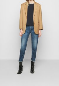 Beige Blazer mit zwei Knöpfen, schwarzem Oberteil, blauen Jeans und schwarzen Stiefeletten mit Absatz; verfügt über eine taillierte Passform und klassische Designelemente.