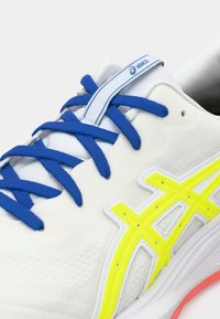 Baskets blanches avec une tige texturée, lacets bleus et accents jaunes vifs. Présente une semelle courbée et un logo sur la languette.