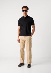 Črna polo majica z diskretnim logotipom, v kombinaciji s svetlim hlačami in temnimi čevlji. Model nosi sončna očala in stoji pred nevtralno ozadjem.