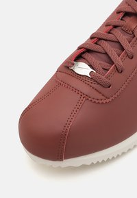 Gros plan d'une basket en cuir bordeaux avec des lacets assortis et une semelle blanche, ornée d'un charm en métal argenté.