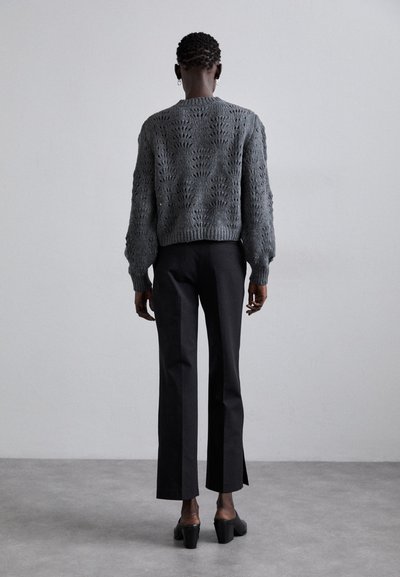 Gray knitted sweater with a detailed openwork pattern, paired with black tailored trousers and heeled shoes, viewed from the back.