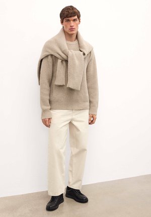 Suéter de punto grueso beige con textura de canalé, bufanda drapeada, pantalones crema y zapatillas negras sobre una superficie lisa.